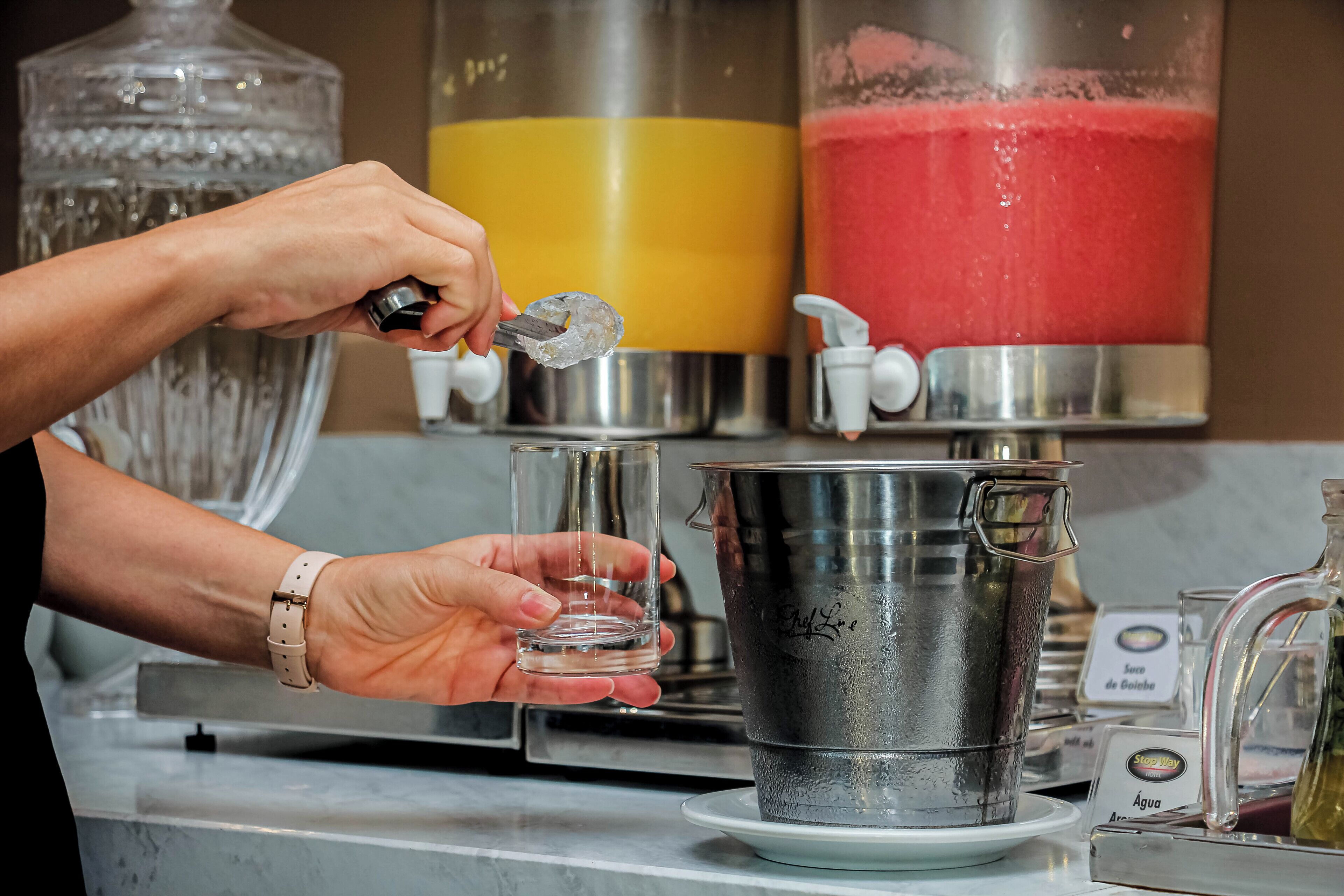A person pouring a drink from a pitcher into a glass. There are two large dispensers in the background, one with a yellow liquid and the other with a red liquid.