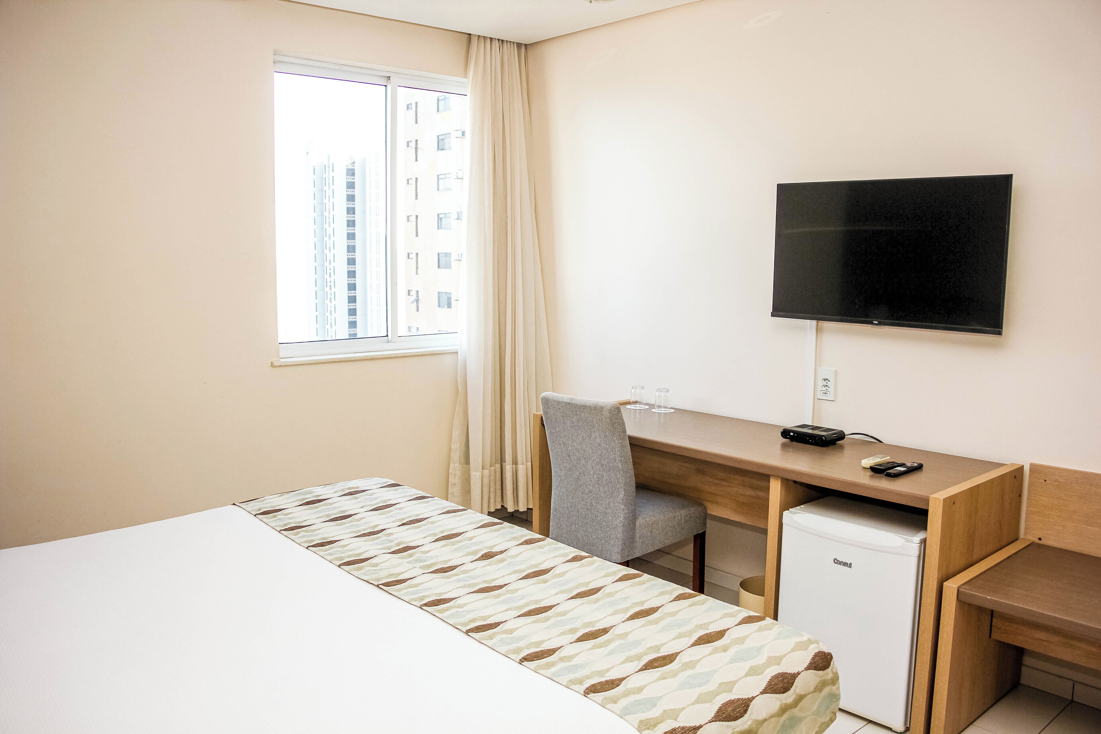 Habitación de hotel con cama, escritorio, computadora, televisor y refrigerador pequeño.