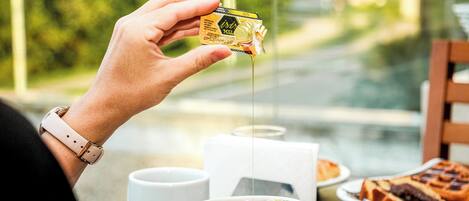 A person holding a small packet of honey, with a cup of tea and a plate of food on a table.