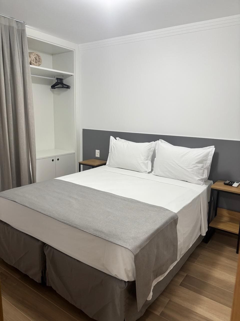 Una cama bien hecha con colcha gris y blanca, una mesita de noche de madera con lámpara y un armario con ropa colgada.
