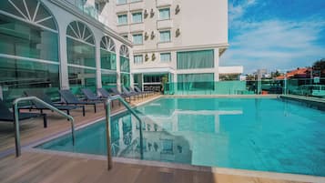 Uma piscina com espreguiçadeiras, um guarda-corpo de vidro e um edifício ao fundo com janelas amplas e varandas.