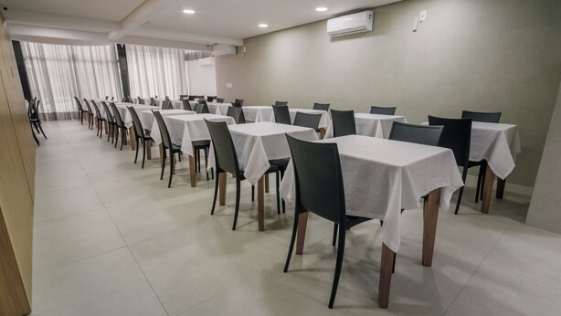 Un salón de conferencias con varias mesas y sillas, unidades de aire acondicionado en la pared, y un ventanal con cortinas.