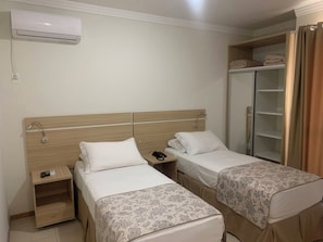 Habitación de hotel con dos camas individuales, cabecero de madera, mesita de noche con teléfono y control remoto, armario y unidad de aire acondicionado.