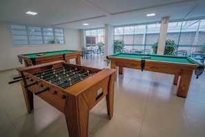 A pool table and a foosball table in a room with large windows and a view of a building outside.