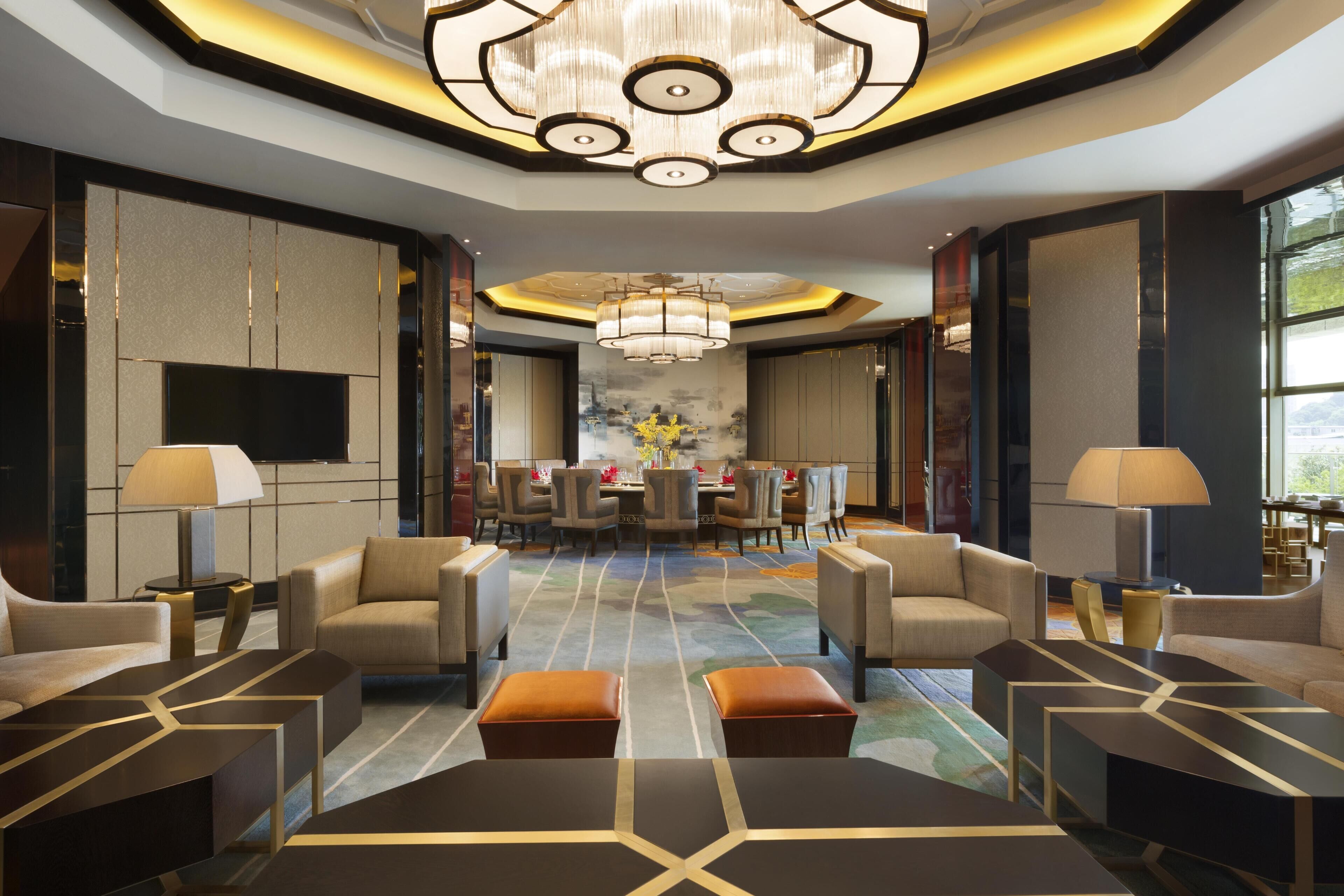 A modern hotel lobby with a central dining area, comfortable seating, and a large chandelier.