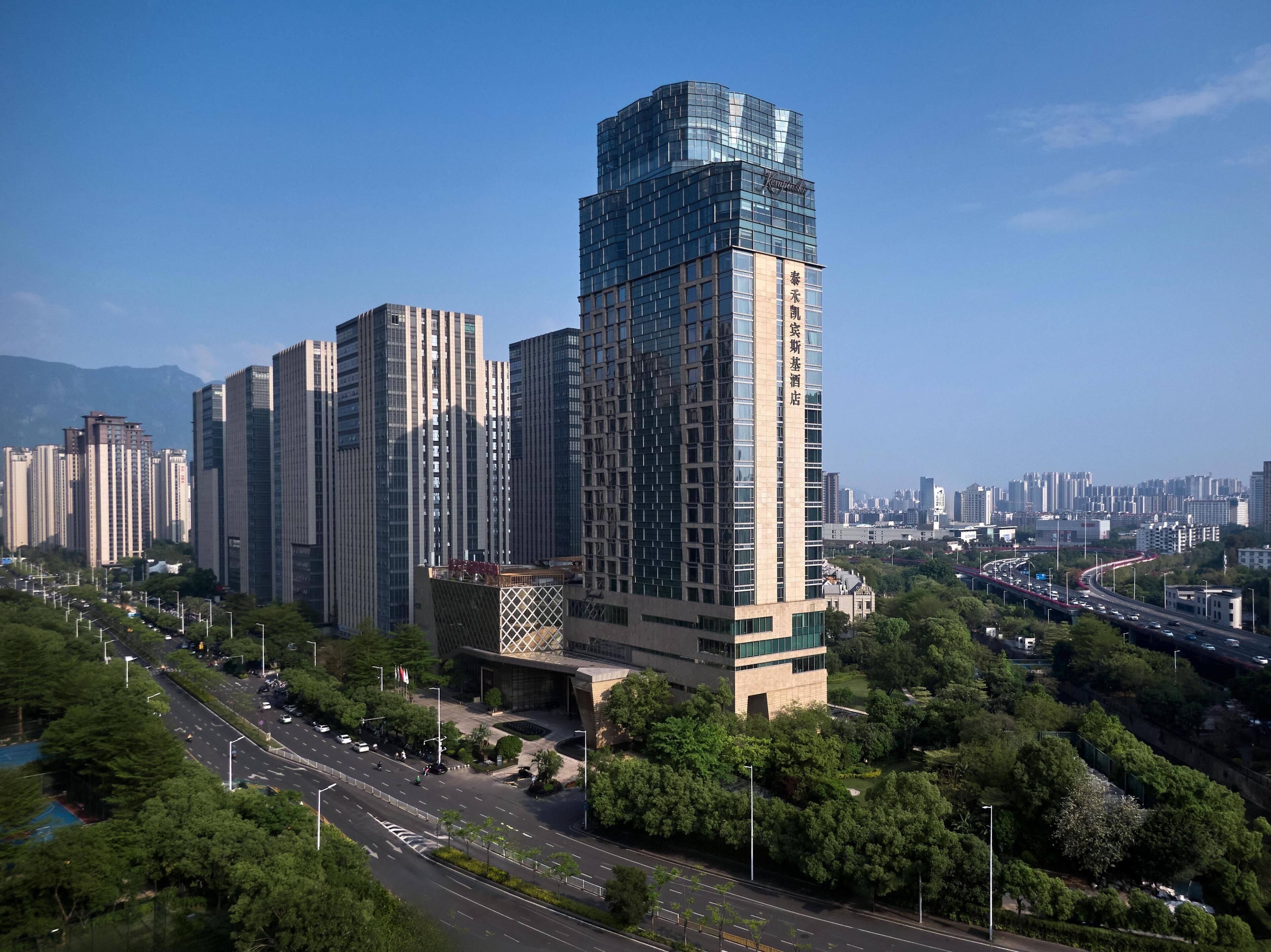 Un edificio alto con fachada de vidrio moderna, rodeado de vegetación y una calle concurrida.