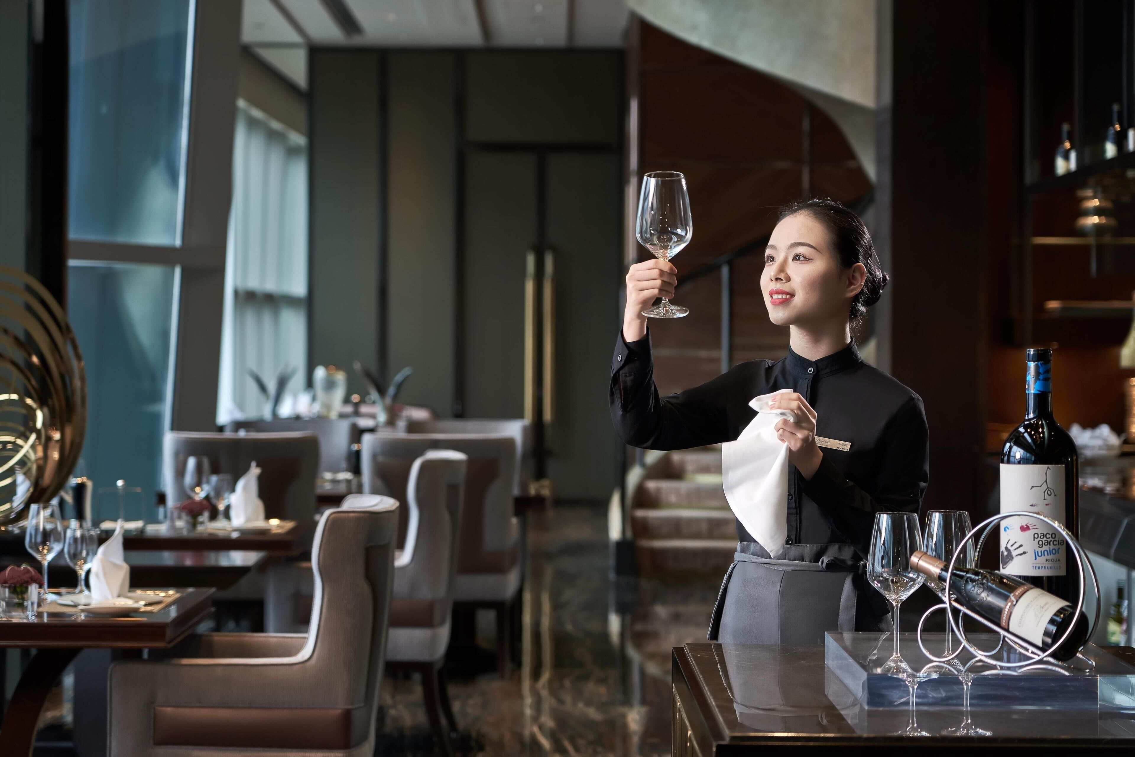 A person holding a wine glass in a restaurant setting with tables, chairs, and a wine bottle on the counter.