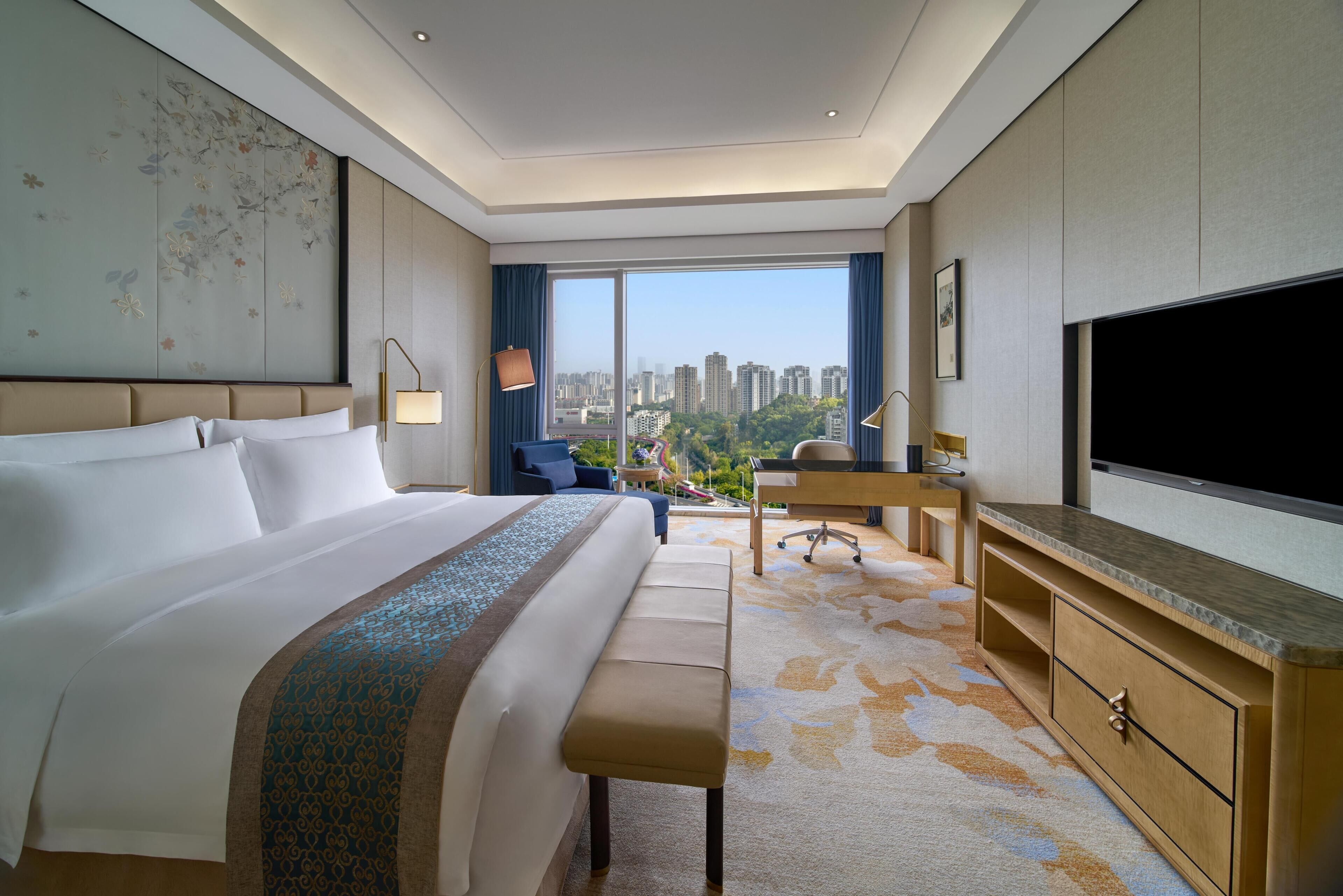 A hotel room with a large bed, a desk, a TV, and a view of the city.