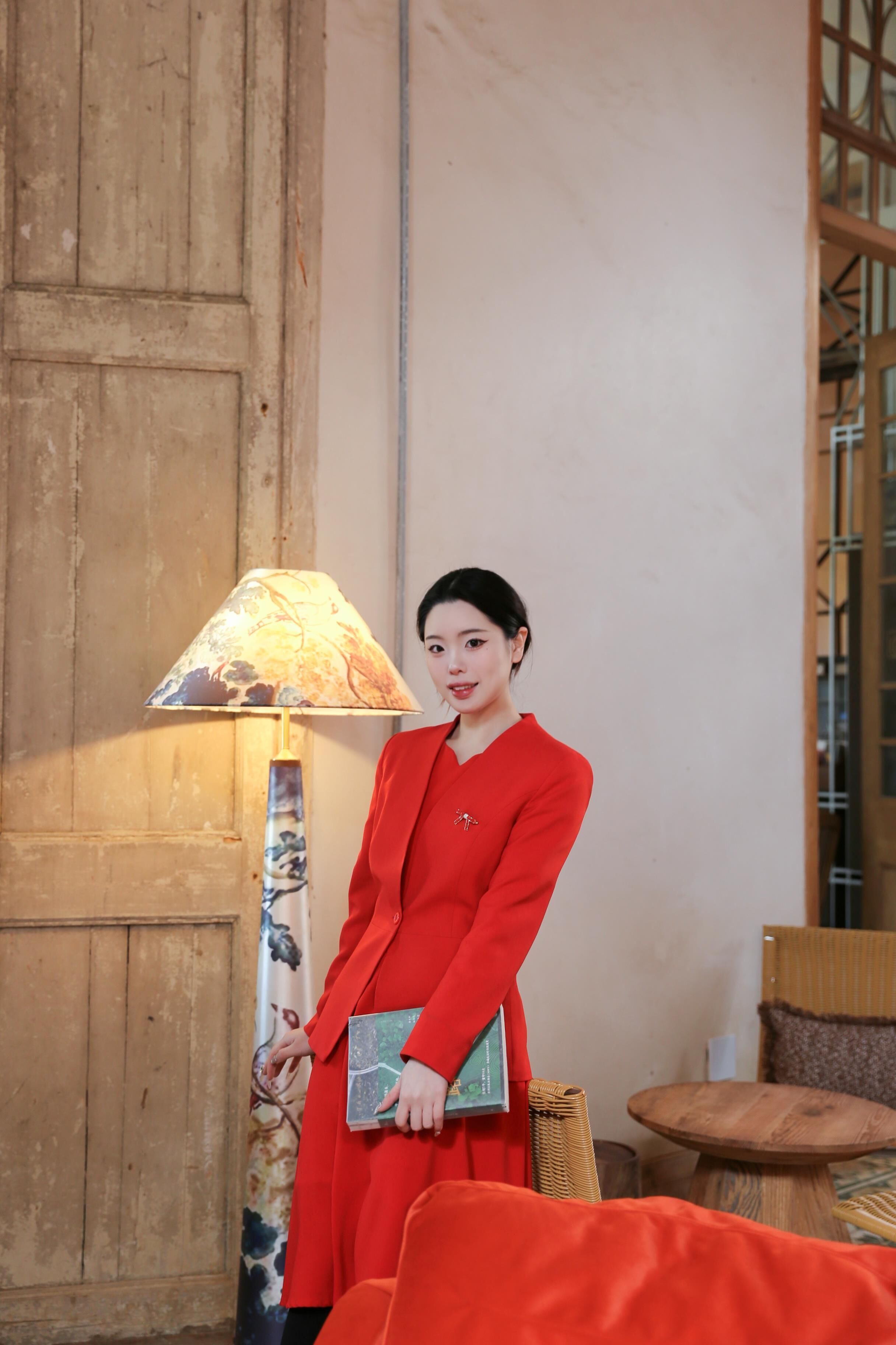 A person in a red outfit standing next to a lamp and a book.