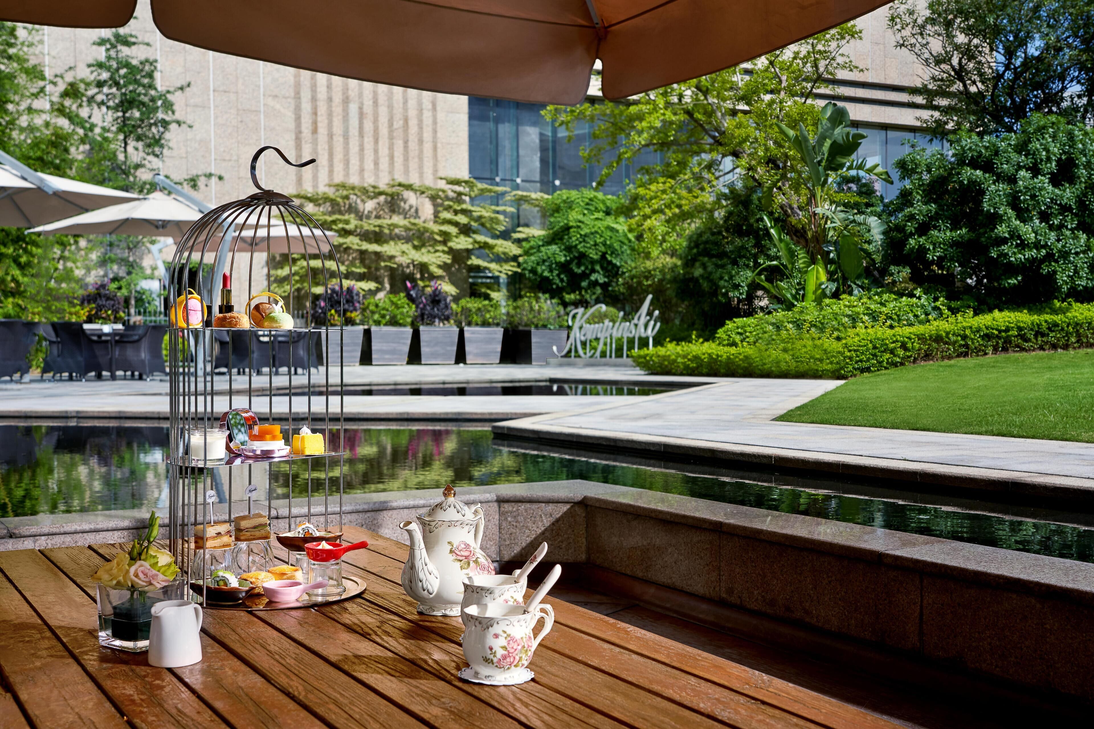 A table set with tea service and a birdcage with decorative items, overlooking a water feature and greenery.
