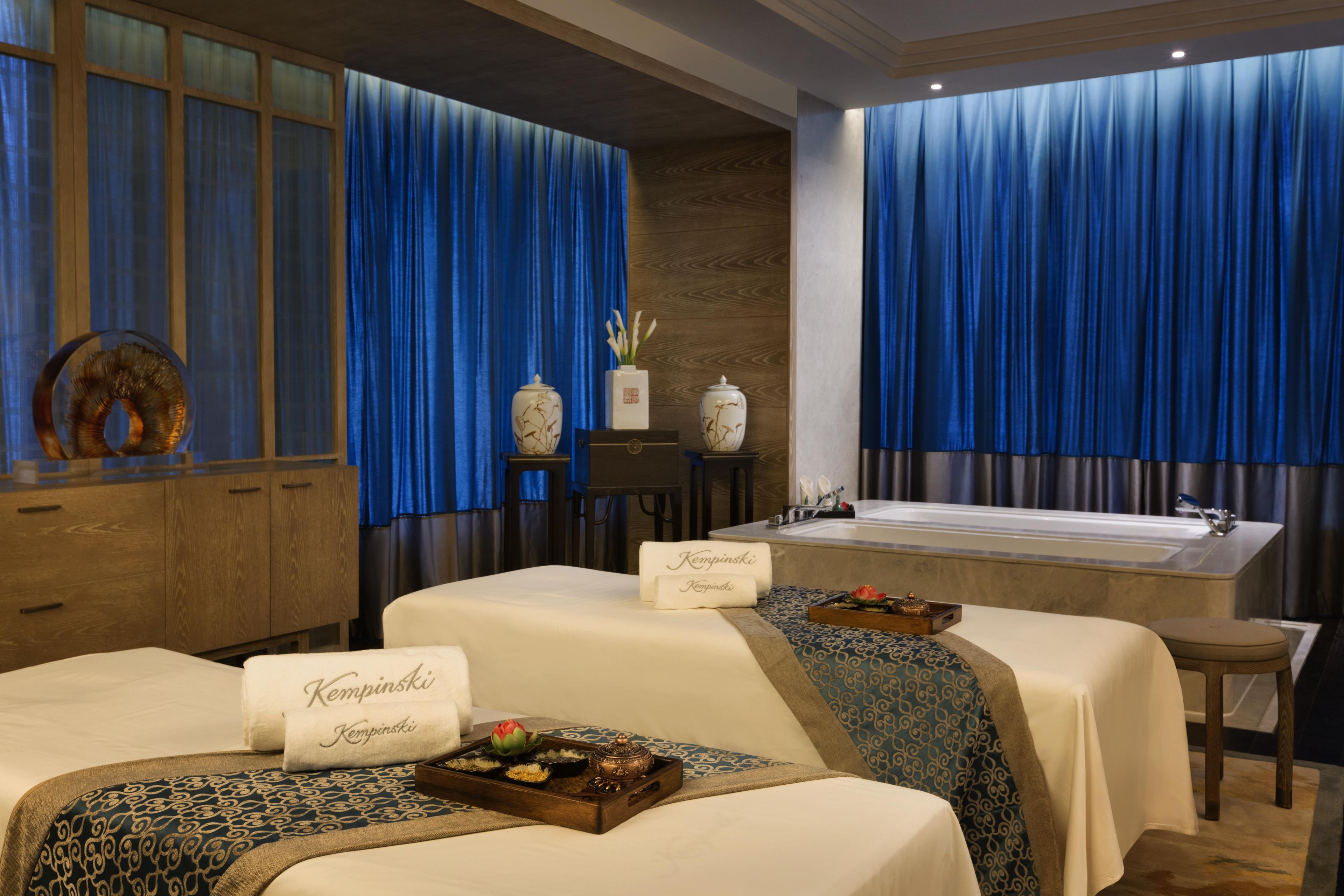 A spa room with two massage tables, a bathtub, and a large window with blue curtains.