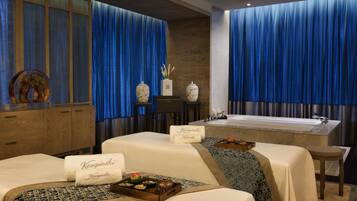 A spa room with two massage tables, a bathtub, and a large window with blue curtains.