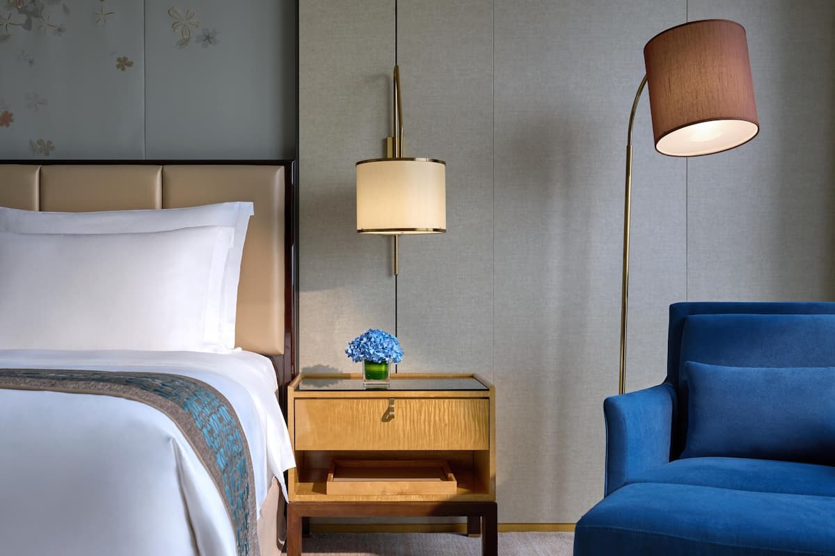 Une chambre d’hôtel avec un lit, une table de chevet ornée d’un vase de fleurs, une lampe et un fauteuil bleu.