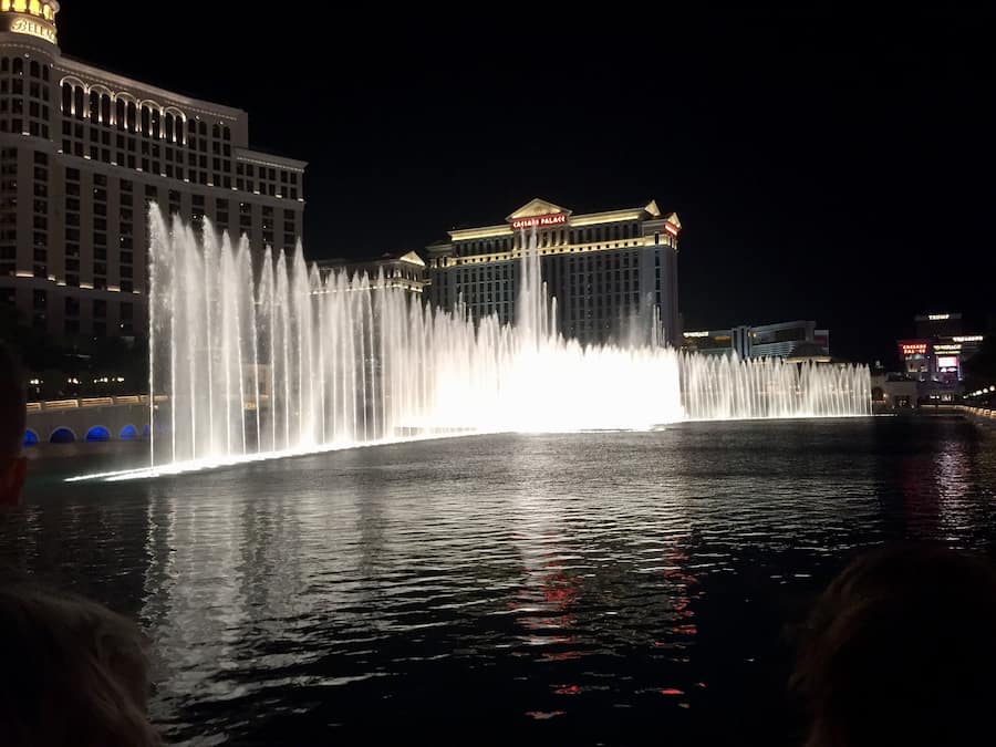 A nighttime view of a large fountain with water jets, surrounded by illuminated buildings including a casino and a hotel.