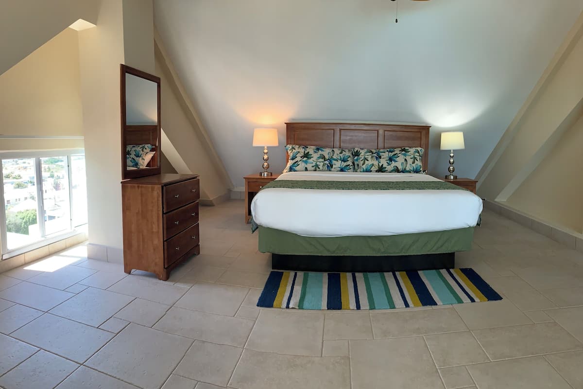 Ein Schlafzimmer mit schrägverdeckter Decke, einem Bett mit grüner Bettdecke, zwei Nachttischlampen, einer hölzernen Kommode, einem Spiegel und einem Streifen Teppich.