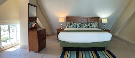A bedroom with a sloped ceiling, a bed with a green bedspread, two bedside lamps, a wooden dresser, a mirror, and a striped rug.