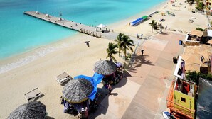 A beach with a pier extending into turquoise waters, a sandy beach with umbrellas, and a hillside with buildings in the background. - Sea Palace (Philipsburg)