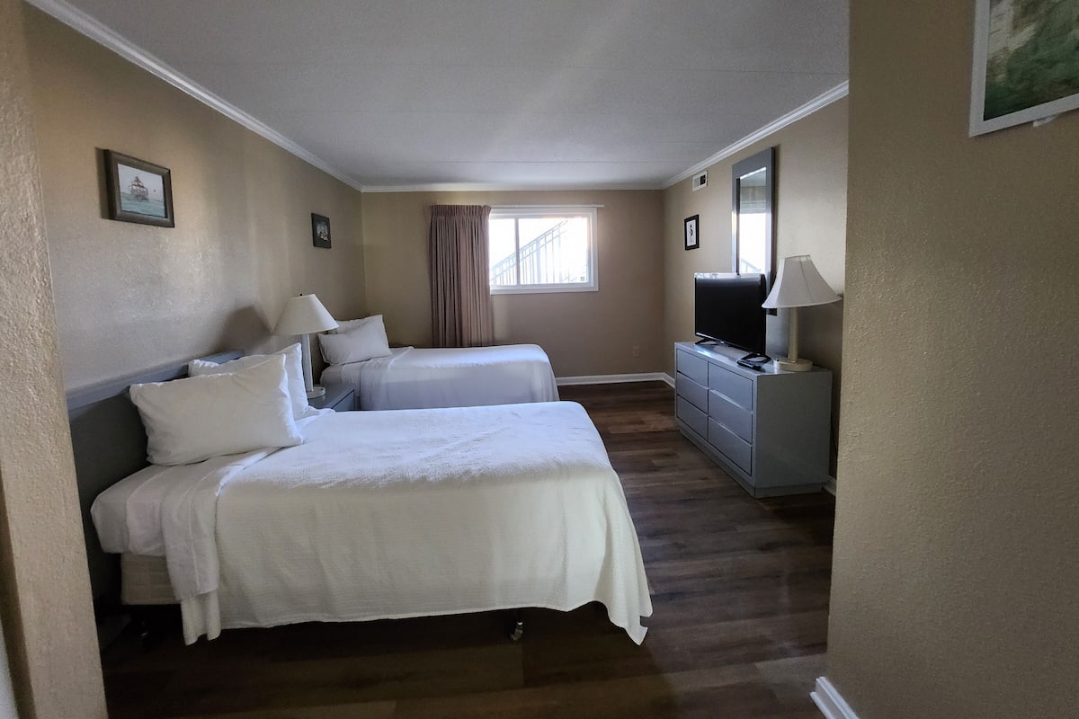 Habitación de hotel con dos camas, un aparador, un televisor y una ventana con cortinas.