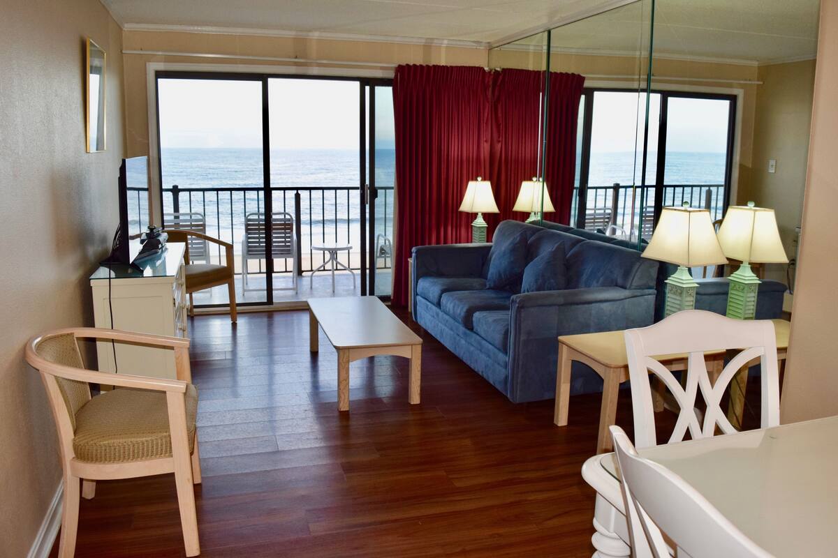 Una habitación con piso de madera, un sofá azul, una mesa blanca y una silla. Tiene un balcón con vista al océano.