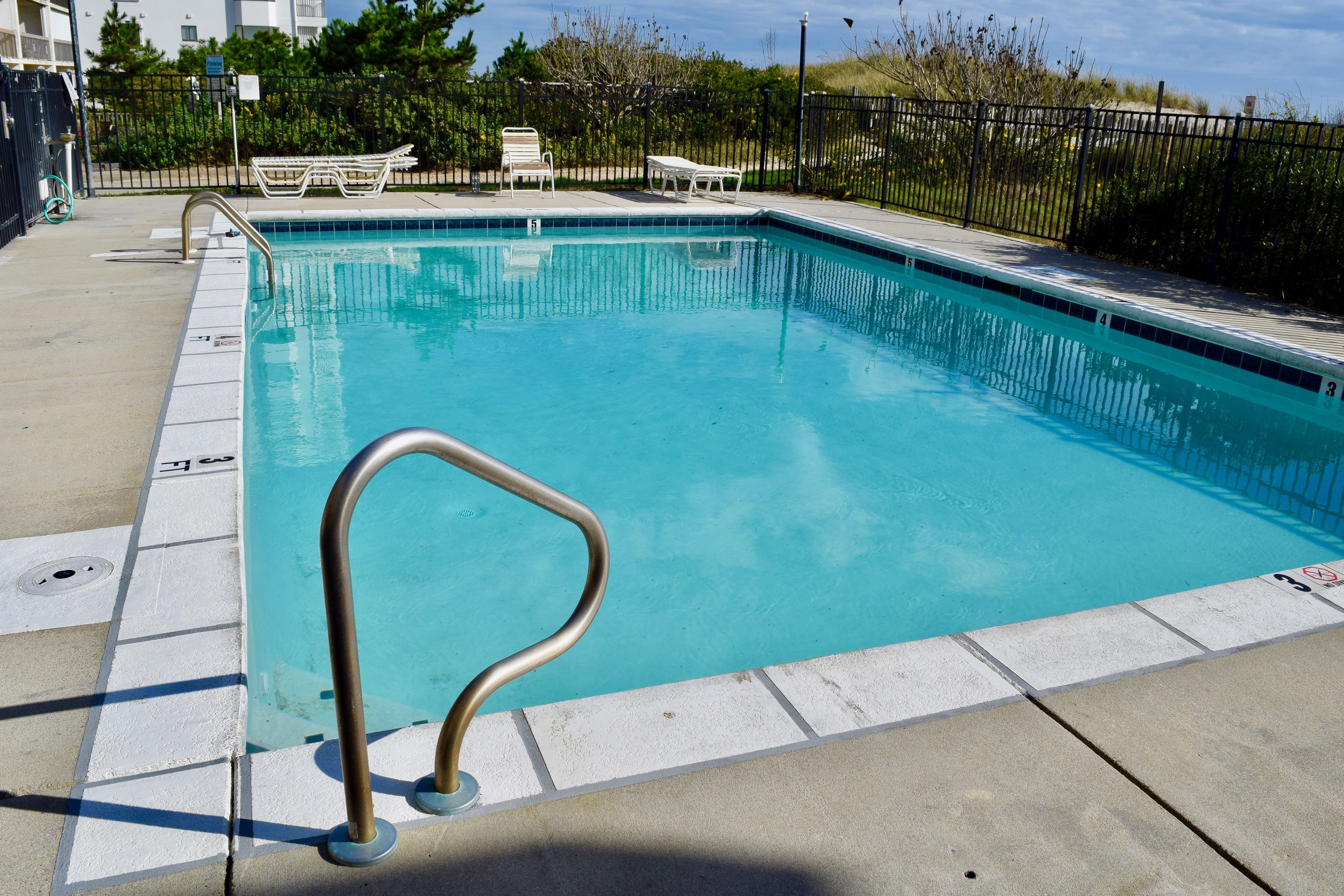 A rectangular swimming pool with a metal handrail on the left side, surrounded by a concrete deck. There are lounge chairs and a small table in the background.