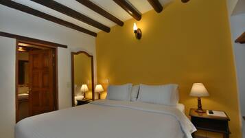 A bedroom with a large bed, wooden beams on the ceiling, a nightstand with a lamp, and a wall-mounted light fixture.