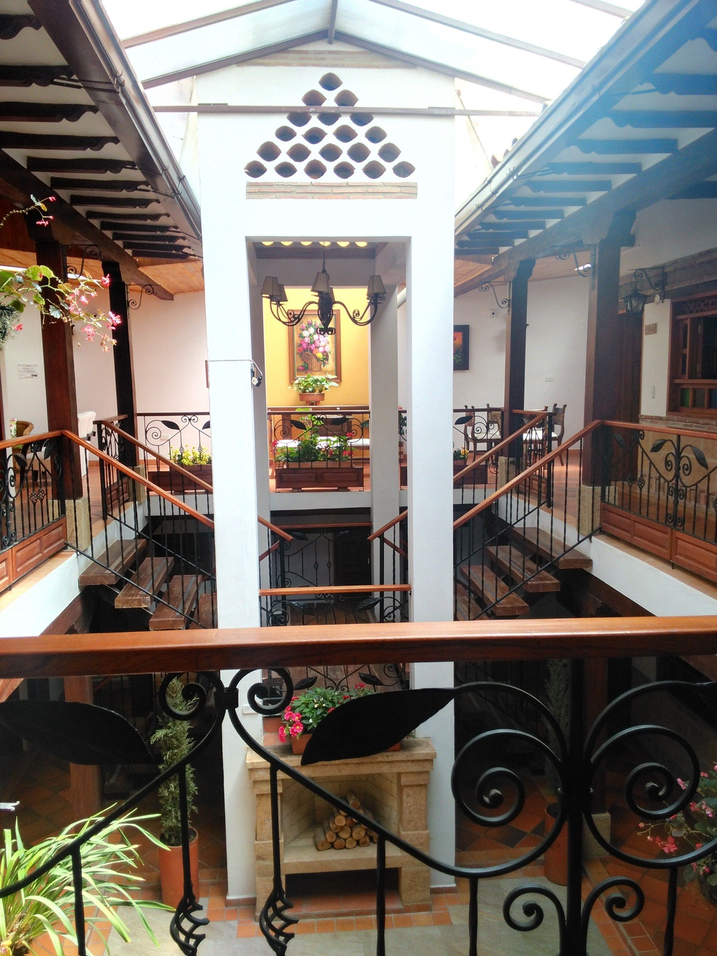 Un interior de varios niveles con una escalera central, barandillas de madera y plantas en macetas.