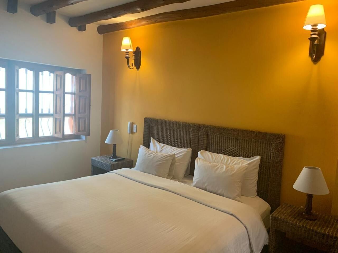 Una cama bien hecha con sábanas blancas, dos lámparas de noche y una ventana con contraventanas de madera.