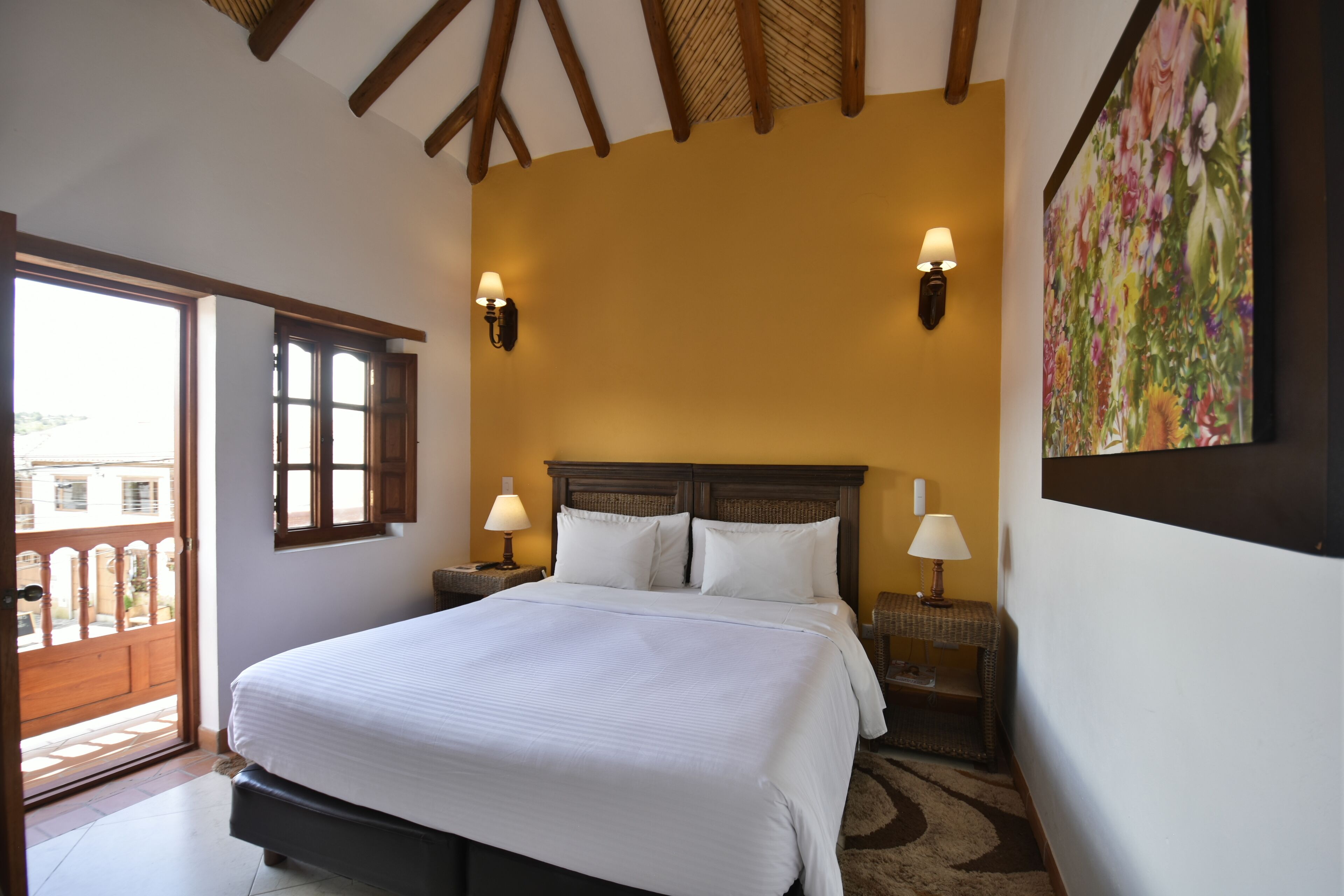 A bedroom with a large bed, two bedside lamps, a wooden headboard, and a floral painting on the wall.