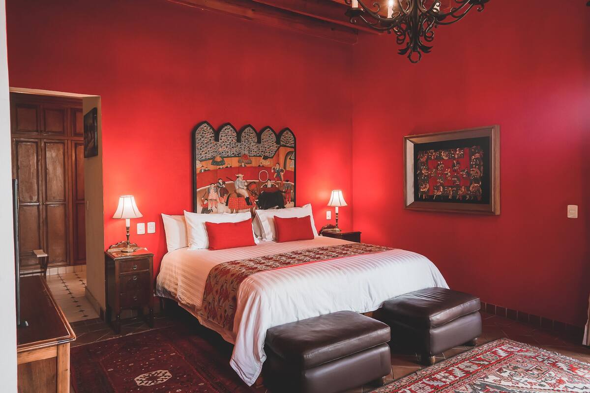 A bedroom with a red wall, a large bed with white bedding, two bedside tables with lamps, a wooden wardrobe, and a patterned rug.