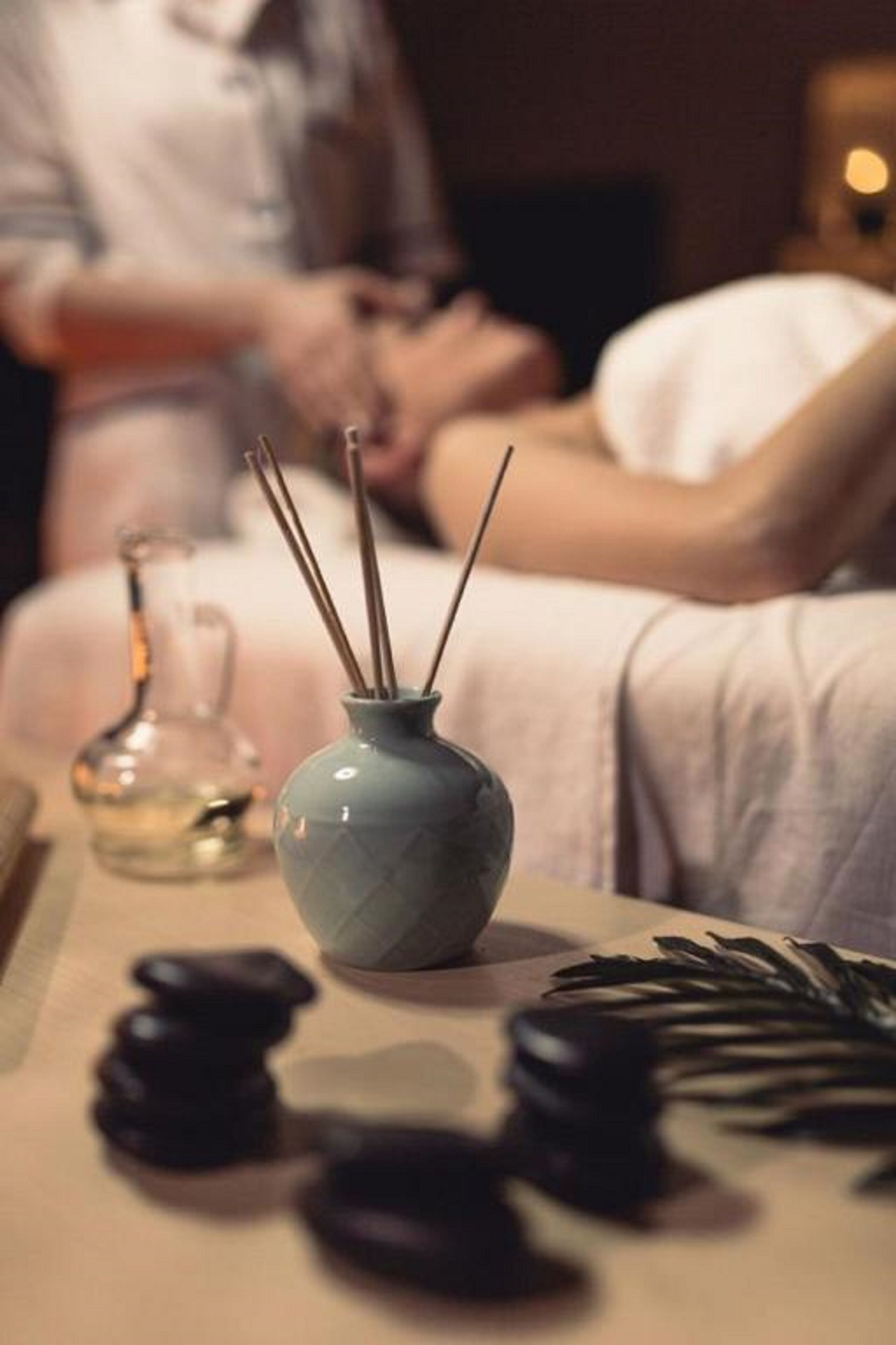 A massage therapy session with a person lying on a massage table, a small blue vase with reed diffusers, and a glass bottle on the table.