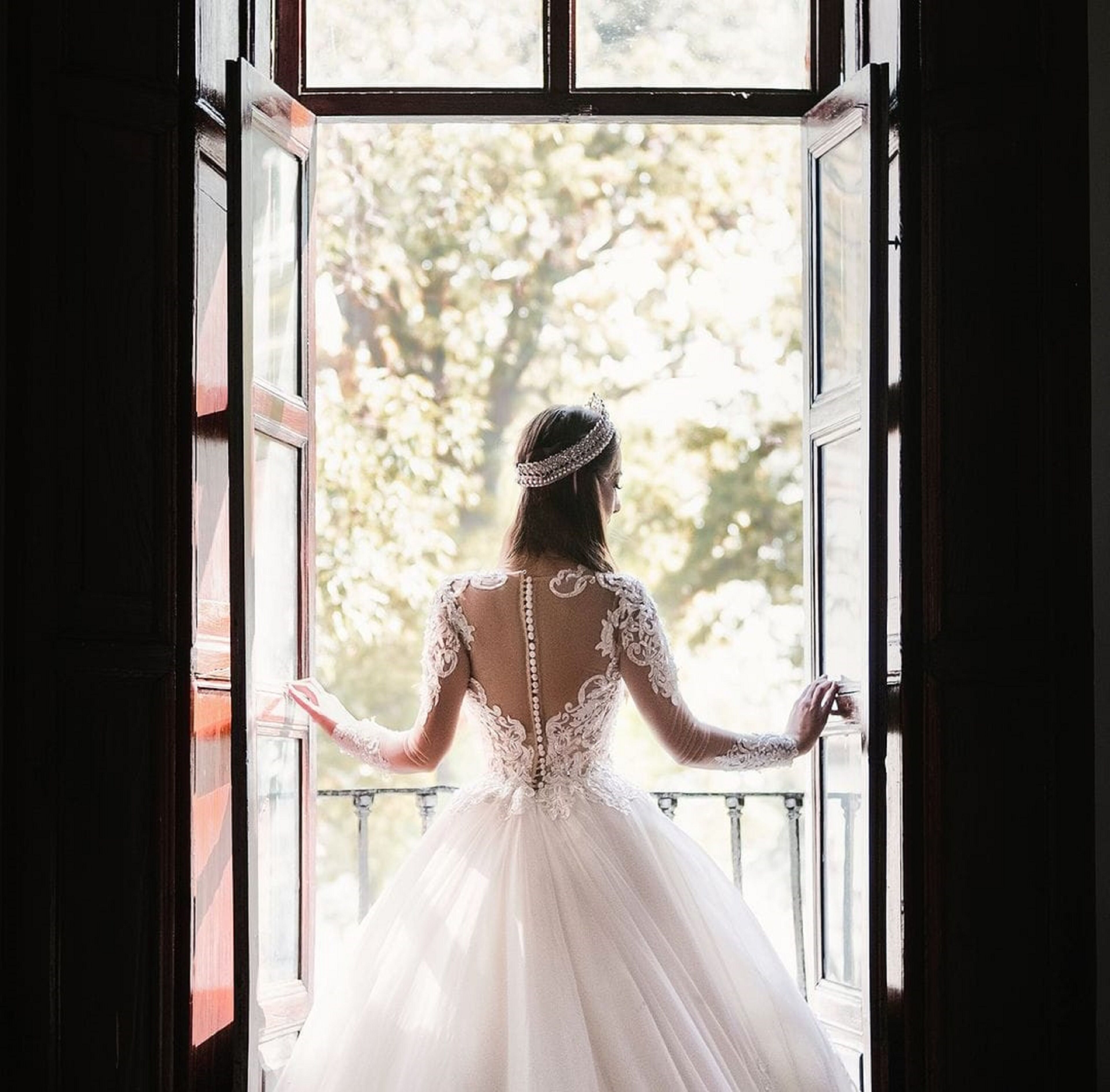 Eine Braut im weißen Hochzeitskleid steht an einem offenen Fenster und blickt hinaus.
