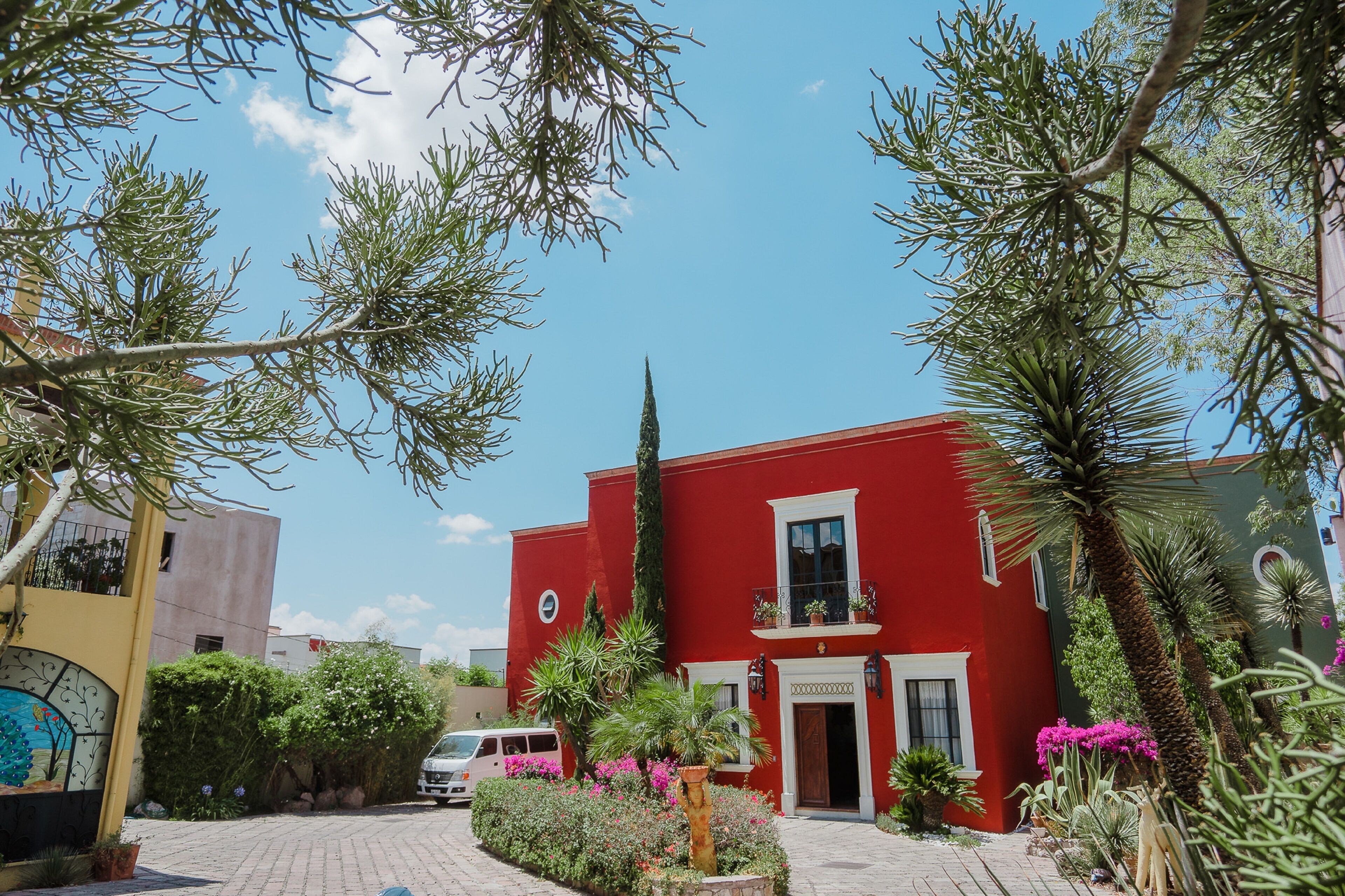 Un edificio rojo con balcón, rodeado de vegetación y un área pavimentada.