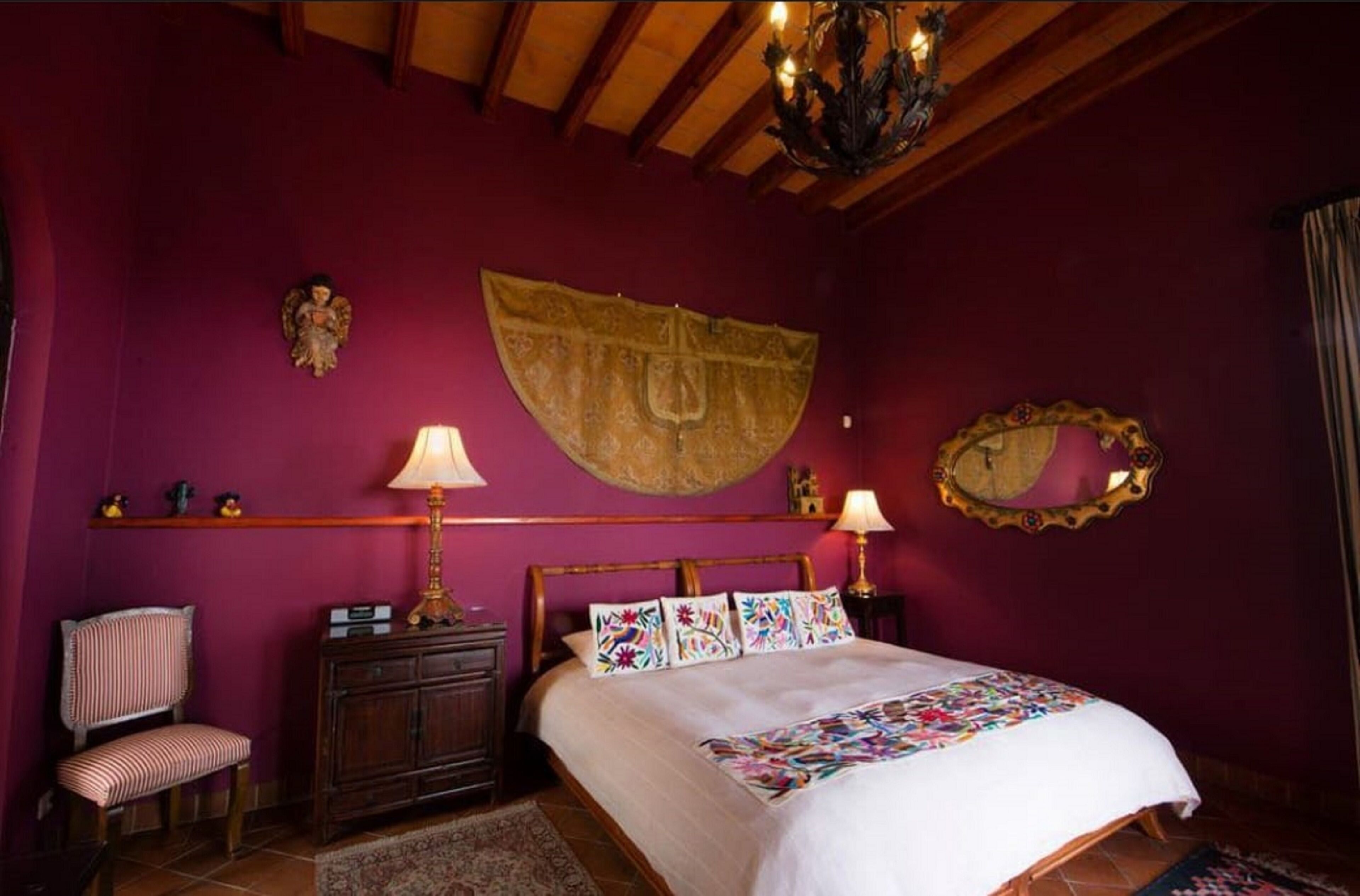 Un dormitorio con cama, dos lámparas de noche, cabecera de madera, un banco, mesita de noche, espejo y un tapiz decorativo.