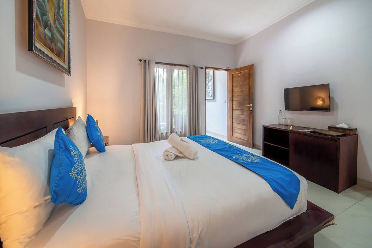 A neatly made bed with blue pillows, a wooden headboard, a blue bedspread, a television on the wall, and a wooden desk with a lamp.