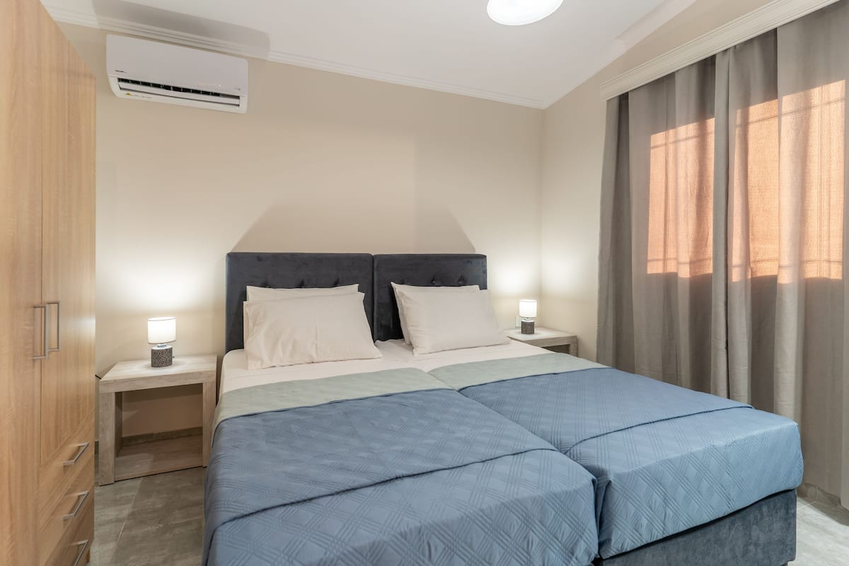 Un lit double avec une couvre-lit bleue, deux tables de chevet avec des lampes, une armoire en bois et une fenêtre avec des rideaux.