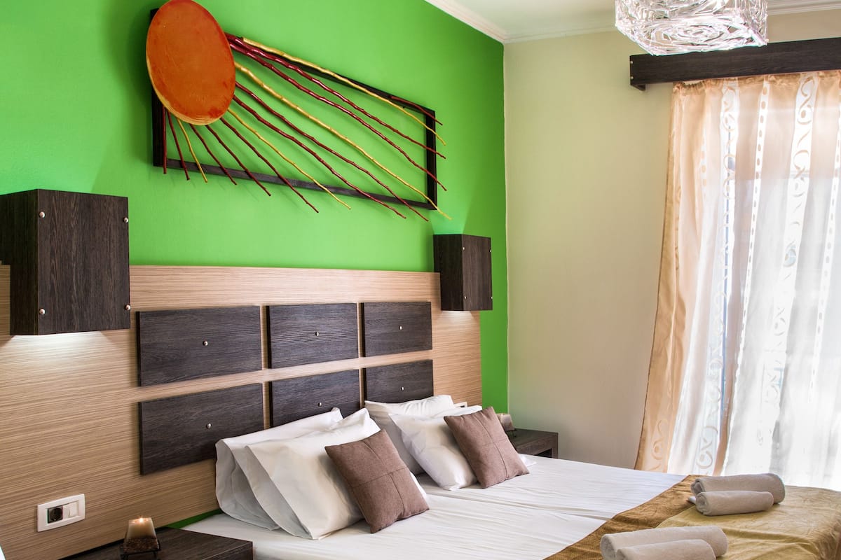 Une chambre d’hôtel avec un mur d’accent vert, une tête de lit en bois, un grand lit avec une literie blanche, deux tables de chevet avec des serviettes et un luminaire suspendu.