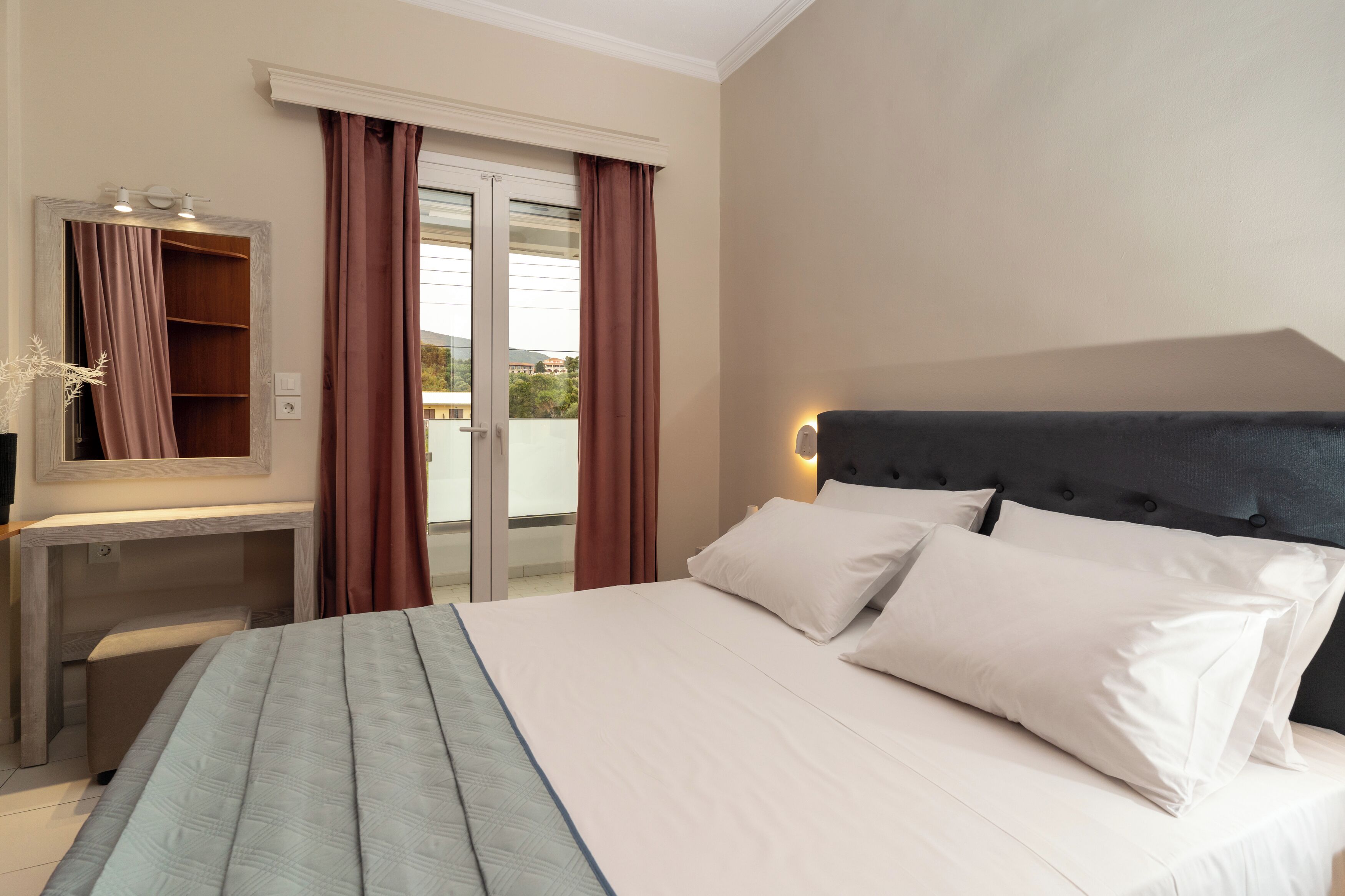 A hotel room with a bed, a desk with a mirror, and a view of the outdoors through a glass door.