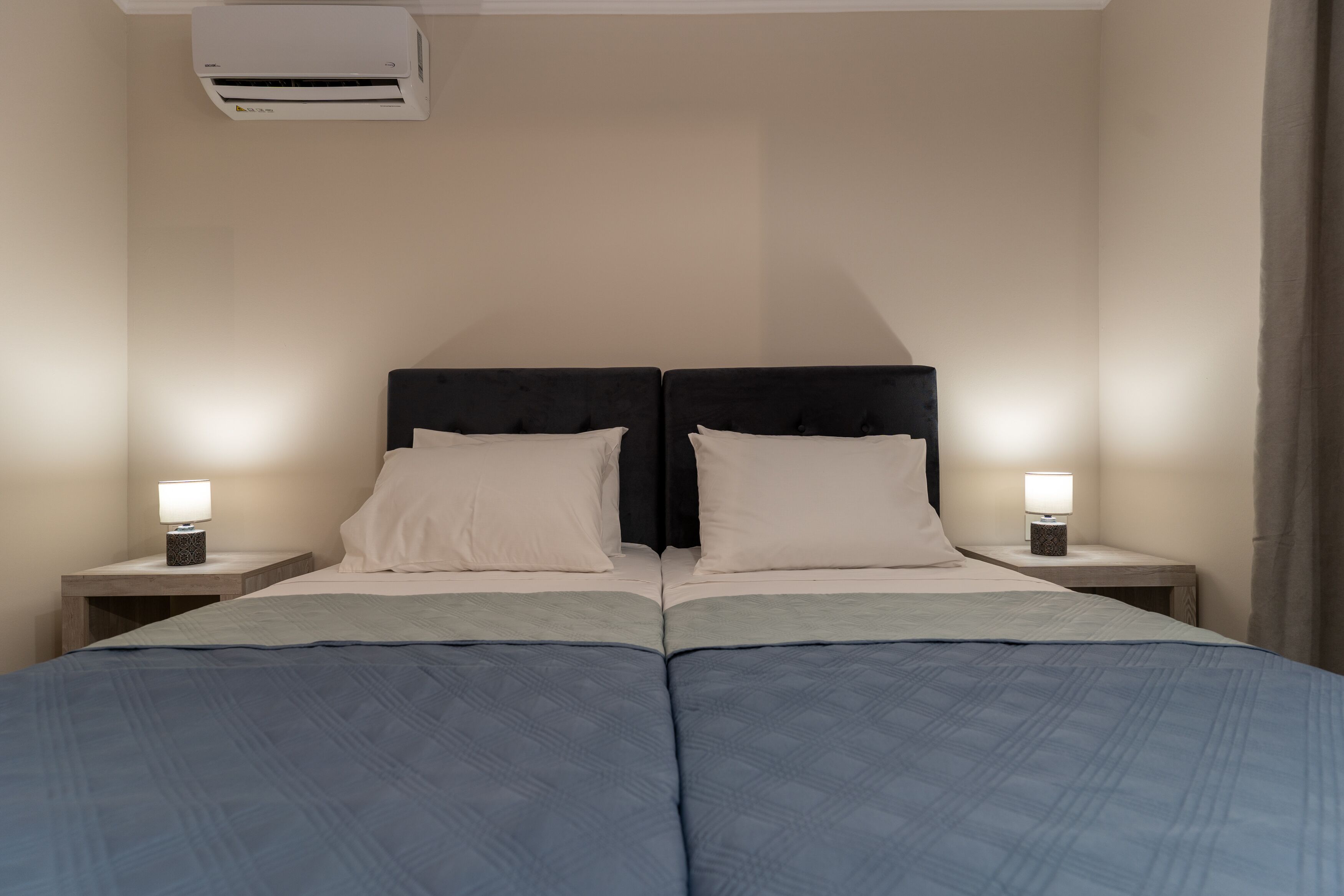 A double bed with a headboard, two bedside tables with lamps, and an air conditioning unit above.