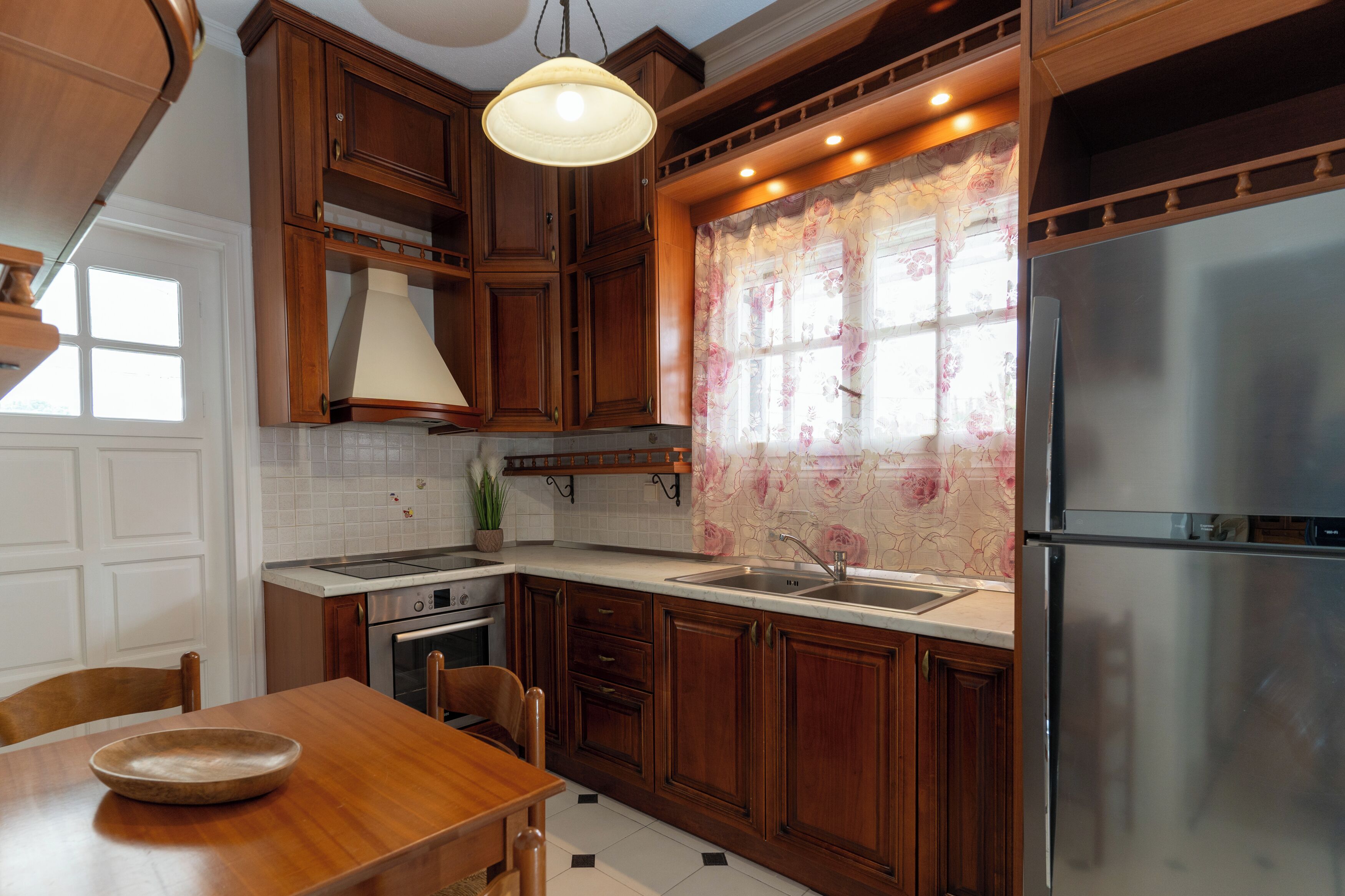 A kitchen with wooden cabinets, a stainless steel refrigerator, a built-in oven, and a window with floral curtains.