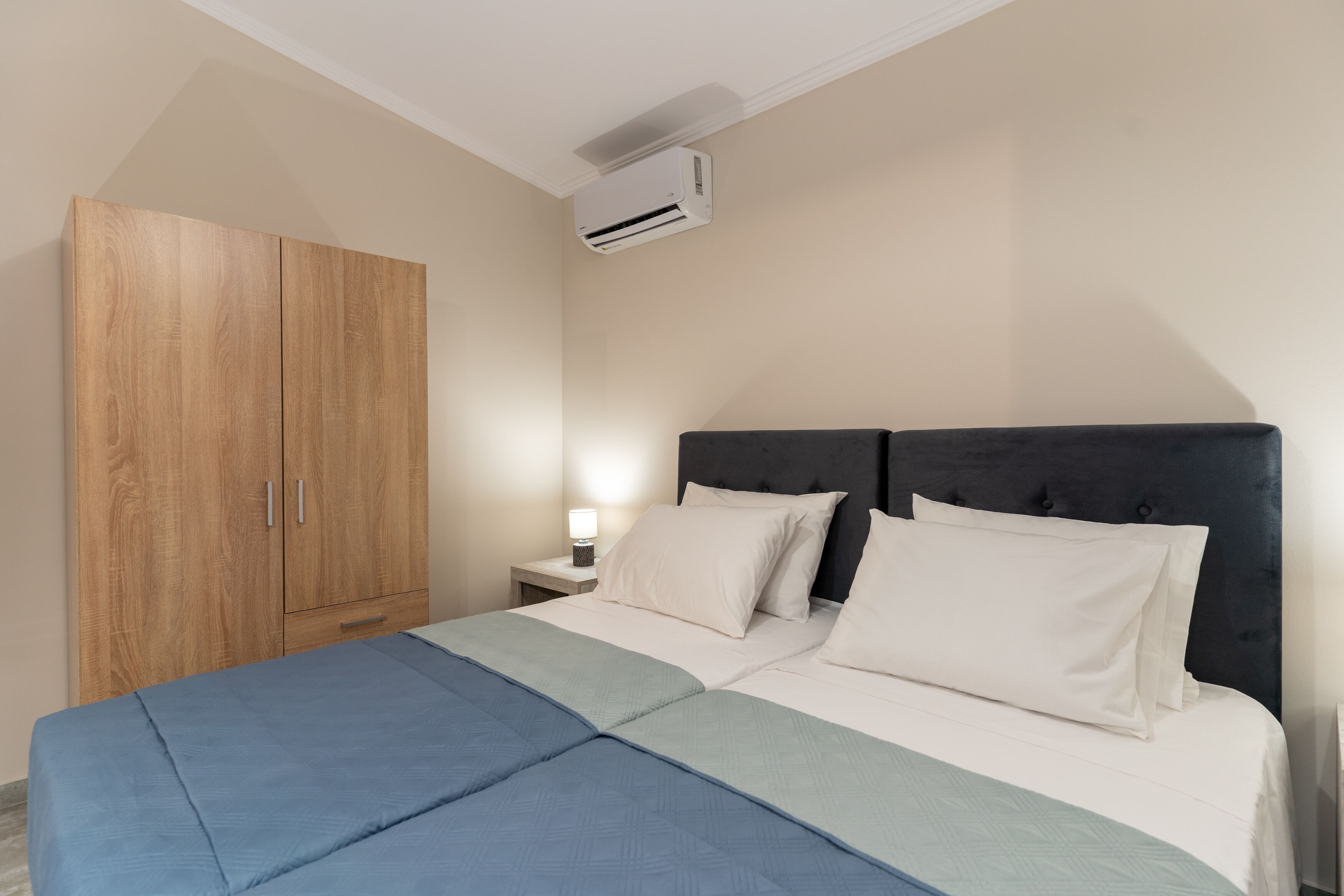 Quarto com cama, guarda-roupa, mesa de cabeceira com abajur e ar condicionado.
