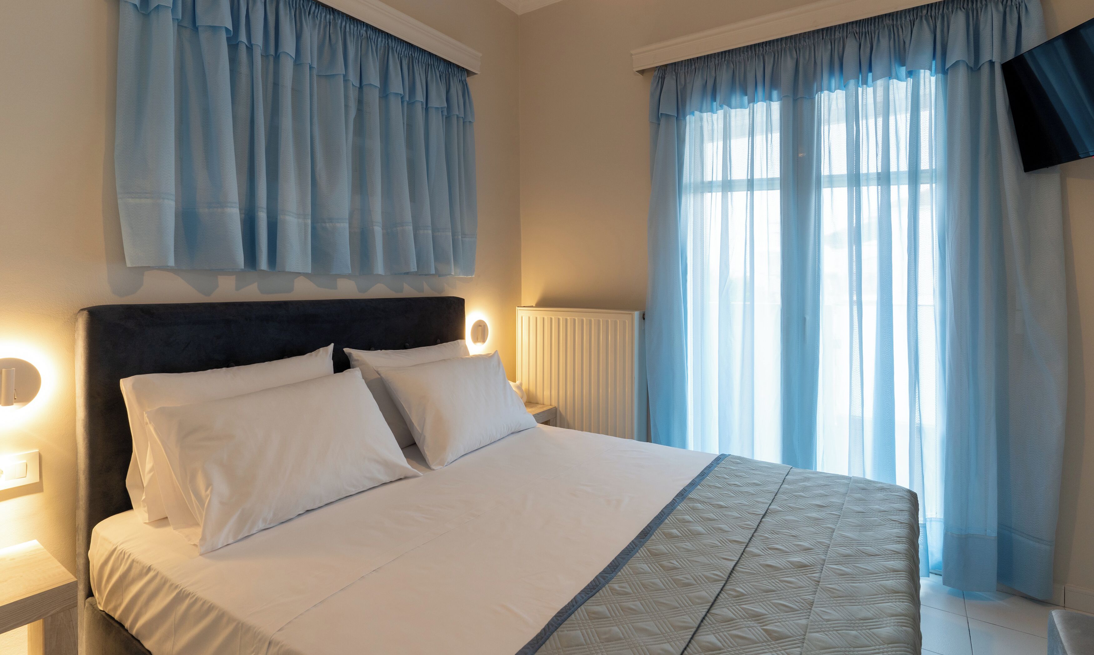 A hotel room with a bed, white bedding, a headboard, bedside lamps, a radiator, and sheer curtains.