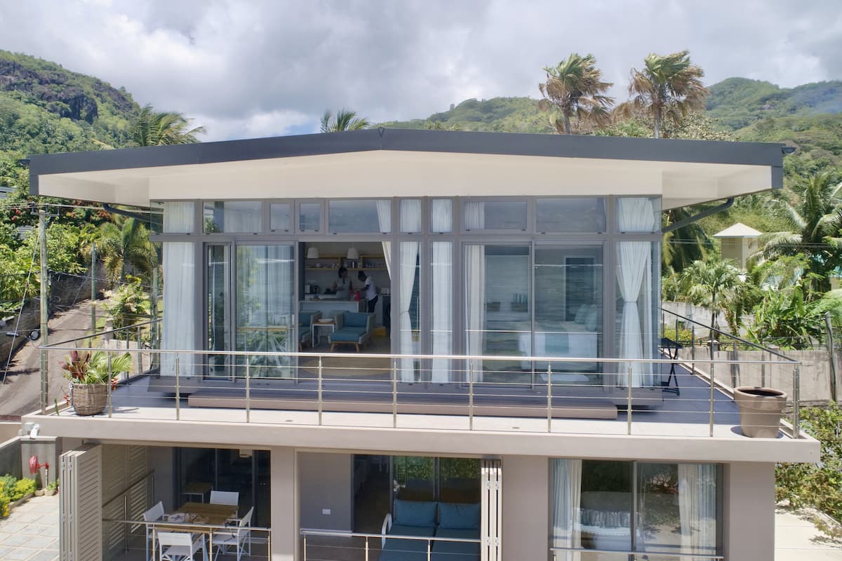 Una casa moderna de dos pisos con grandes ventanales, un balcón y vistas de abundante vegetación y colinas.