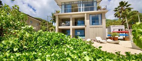 A modern two-story building with balconies, surrounded by lush greenery and palm trees.