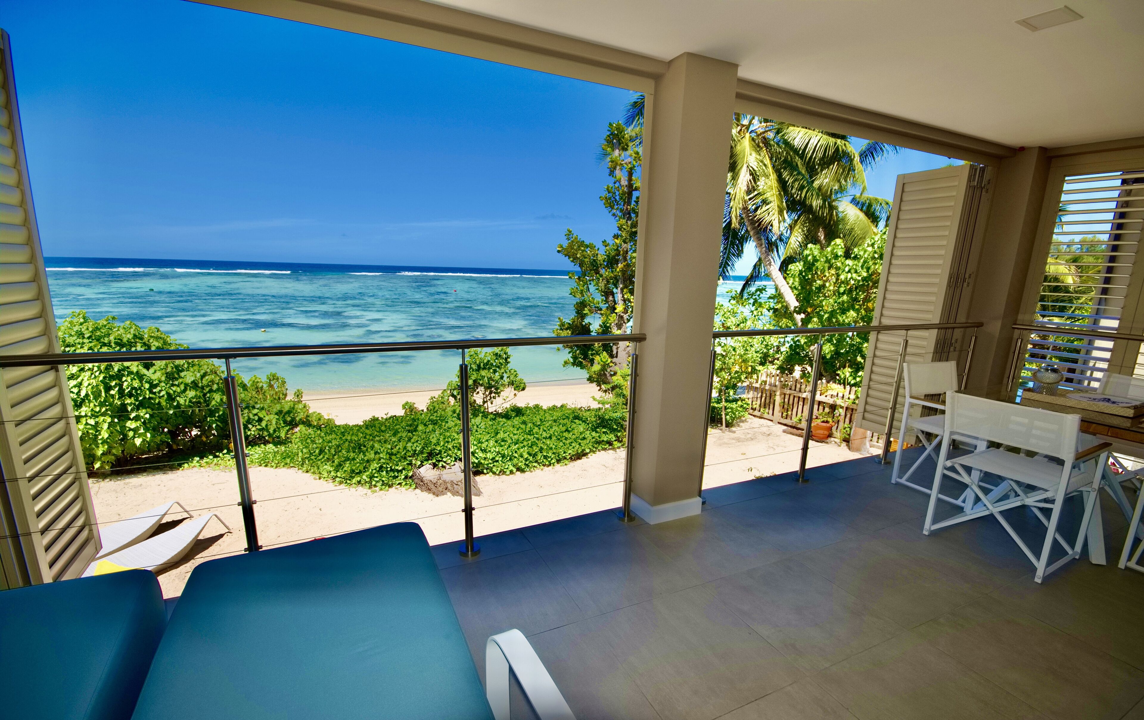 Ein Balkon mit Blick auf den Ozean und den Strand, mit einem blauen Liegestuhl, einem weißen Tisch und einem Blumentopf.
