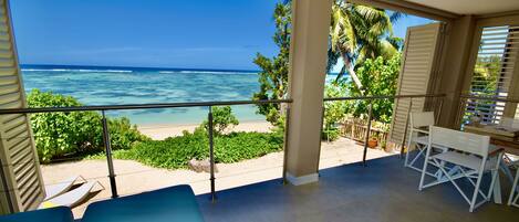 Un balcon avec vue sur l’océan et la plage, comprenant un fauteuil bleu, une table blanche et un pot de fleurs.