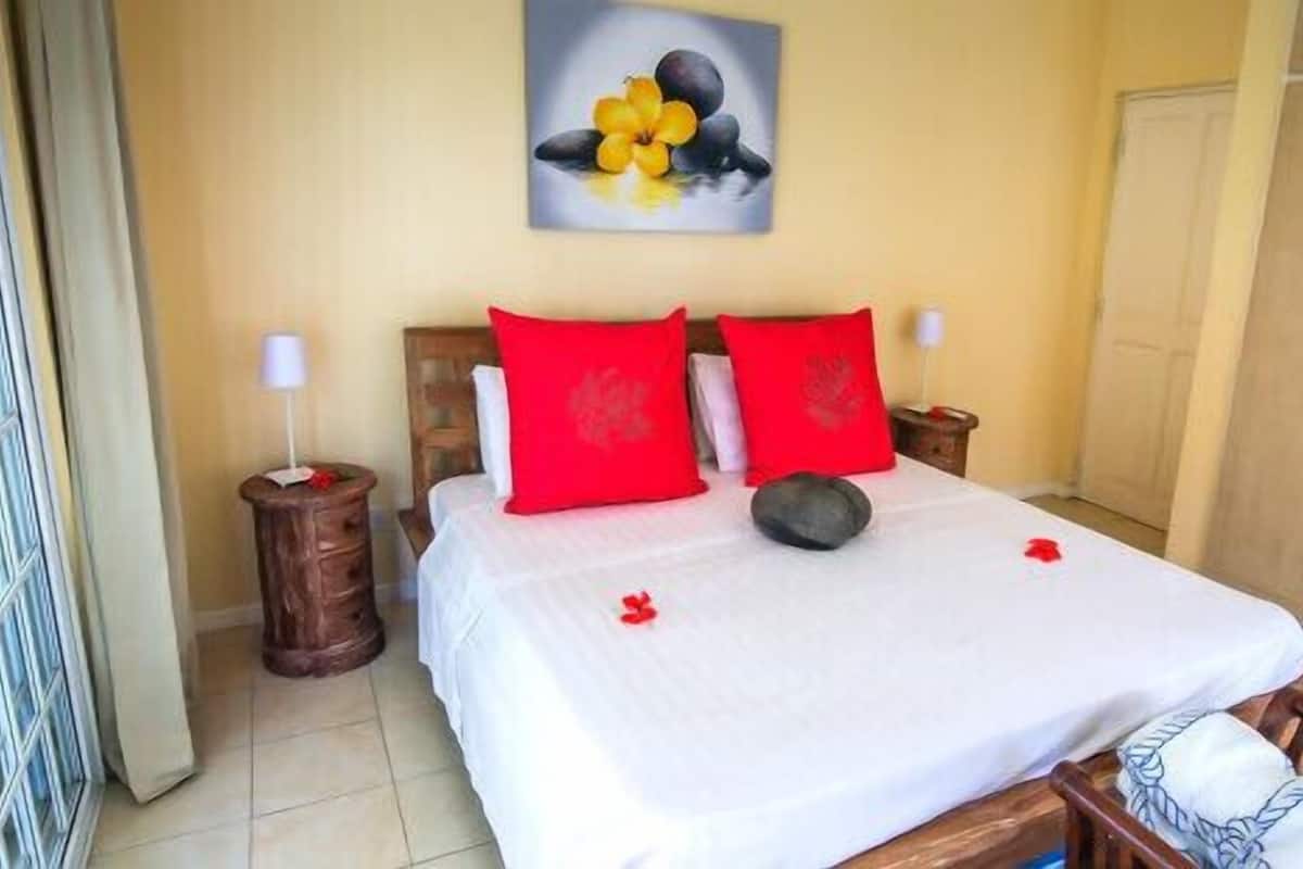 Una habitación de hotel con una cama, dos almohadas rojas, un cabecero de madera, una mesita de noche con una lámpara y un cuadro en la pared.