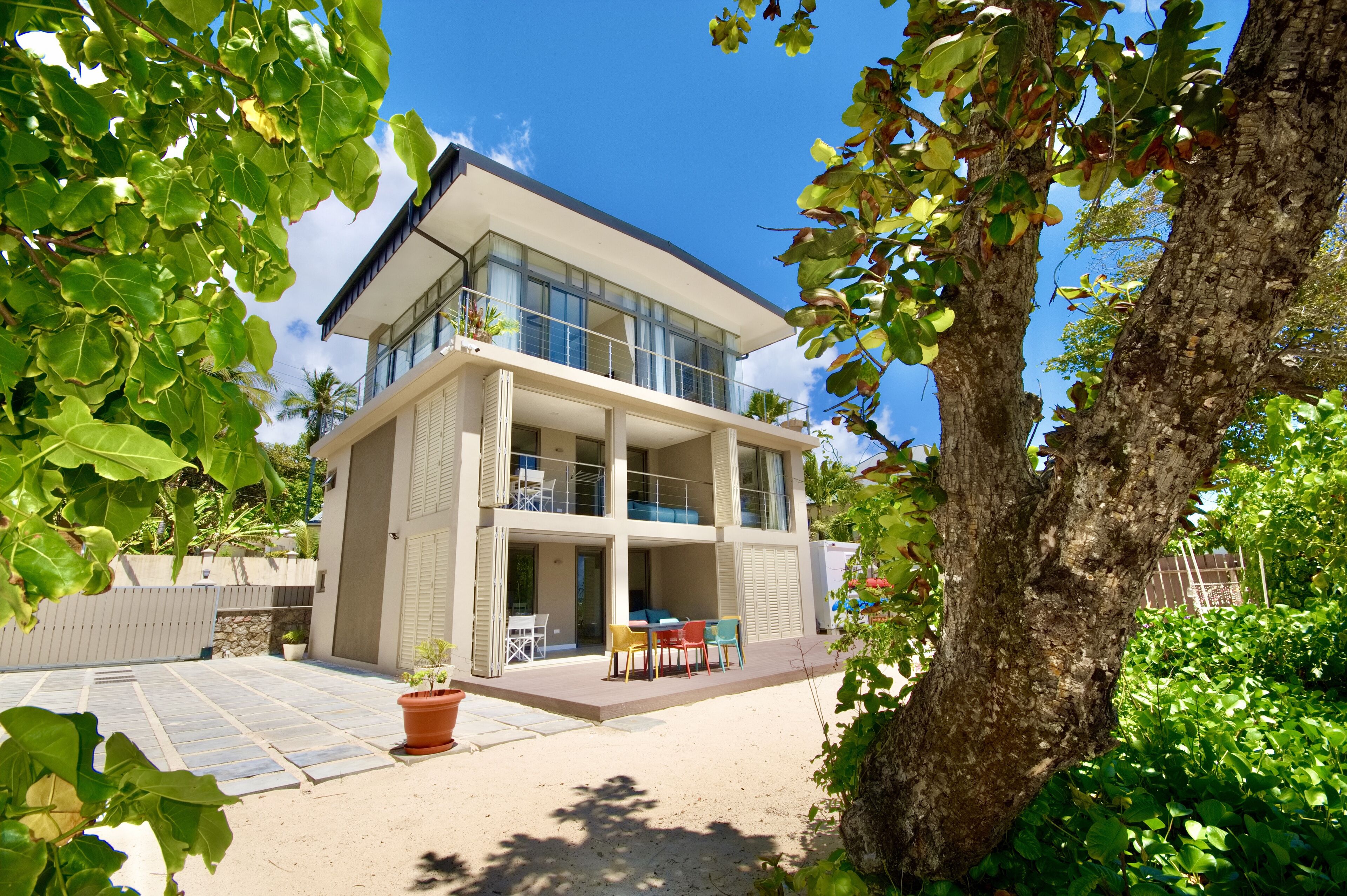 Una casa moderna de dos pisos con un balcón, un patio con mobiliario exterior y un árbol en primer plano.
