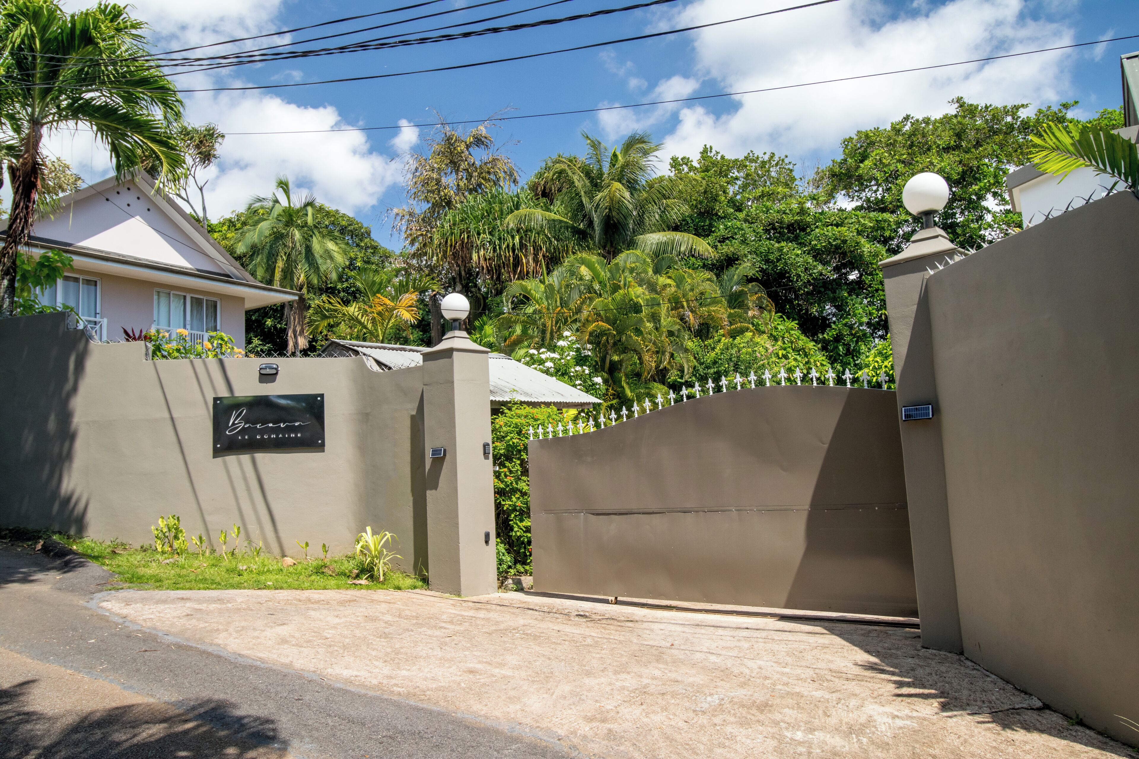 A gated entrance with a sign, a paved driveway, and lush greenery in the background.