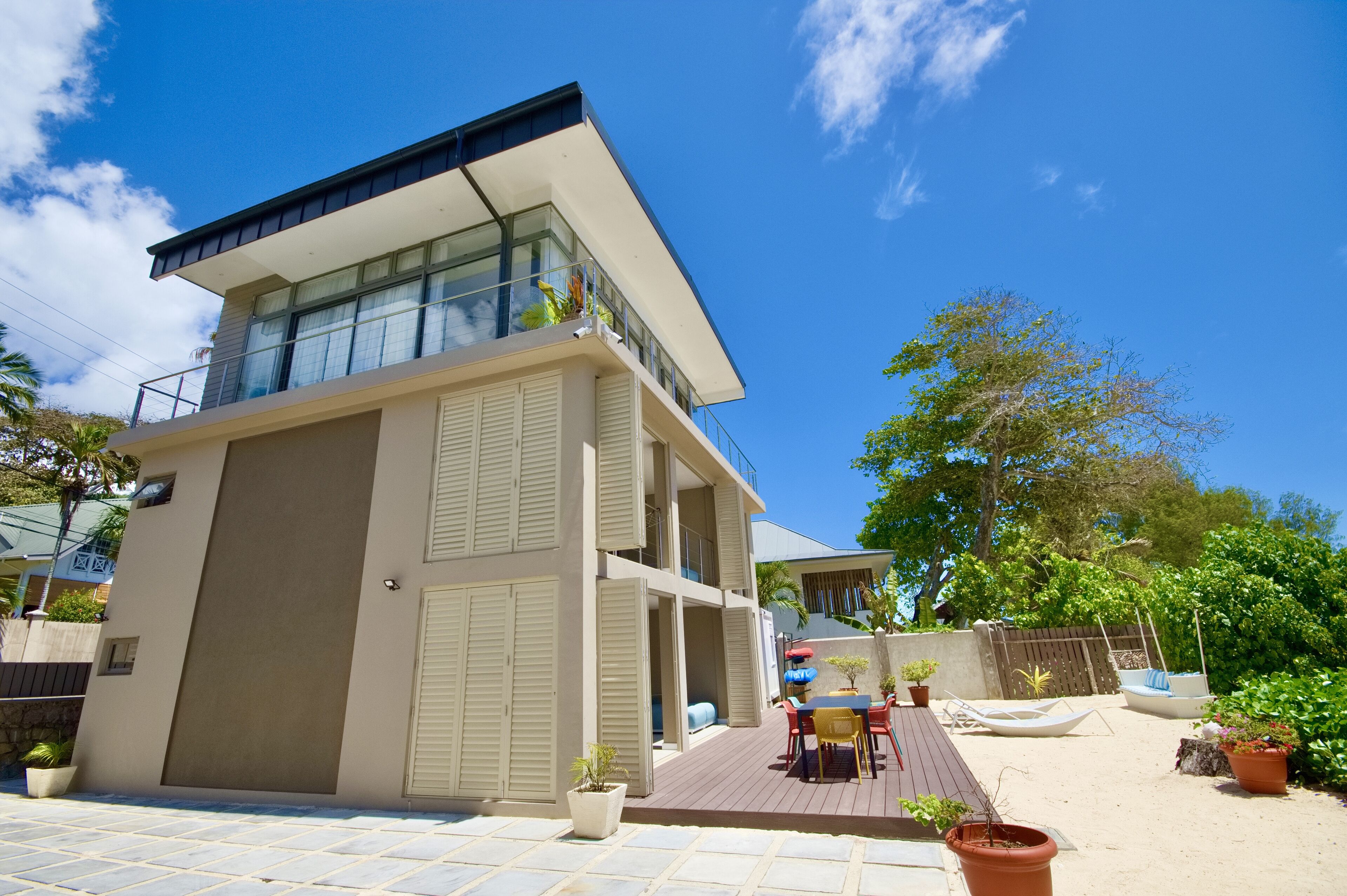Una casa moderna de dos pisos con un balcón, un patio con mobiliario exterior y plantas en macetas.