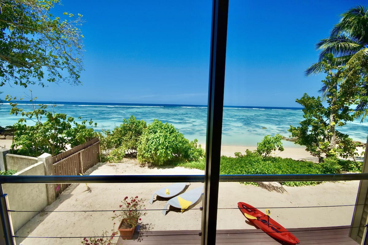 Vista de una playa desde un balcón con baranda de madera, un kayak rojo y sillas de descanso.